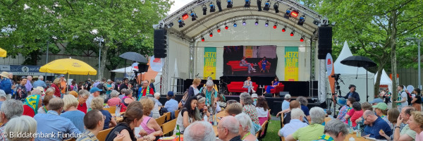 Viele Menschen sitzen vor einer Bühne des Kirchentags in einer Grünanlage zusammen