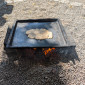 Eine flache, dunkle Metallplatte mit einem kleinen, flachen Stück Teig (Fladen) liegt auf einem niedrigen Feuer. Das Feuer brennt auf einem Sockel aus zwei großen Holzblöcken, alles auf einem kiesigen Untergrund.