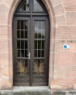 Ein gotisches Steintor mit einer dunklen Holztür, die von einem steinernen Bogen überragt wird, dominiert die Ansicht. Rechts neben der Tür ist ein kleines, rechteckiges Schild mit einem blauen Hintergrund und einem weißen stilisiertem Ohr, welches auf eine vorhandene induktive Höranlage hinweist, angebracht. 