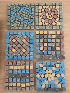 Sechs quadratische Mosaikplatten mit verschiedenen Mustern aus kleinen bunten Steinen auf hellem Untergrund. 