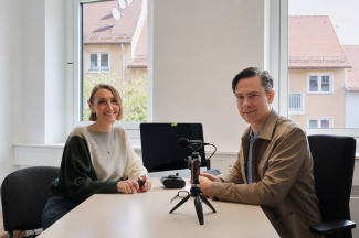 Eine Frau und ein Mann sitzen in einem hellen Büroraum gemeinsam an einem Tisch und lächeln in die Kamera. Zwischen ihnen stehen ein Mikrofon auf einem kleinen Stativ sowie weitere Aufnahmegeräte.